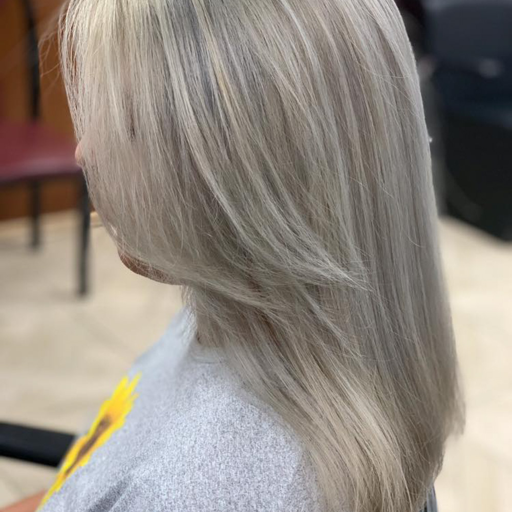 A side profile of a client with medium-length hair colored in a bright, cool-toned platinum blonde or silver hue. The hair is styled with soft, face-framing layers and a smooth, straight blowout, showcasing a seamless transition from root to tip without any brassiness.