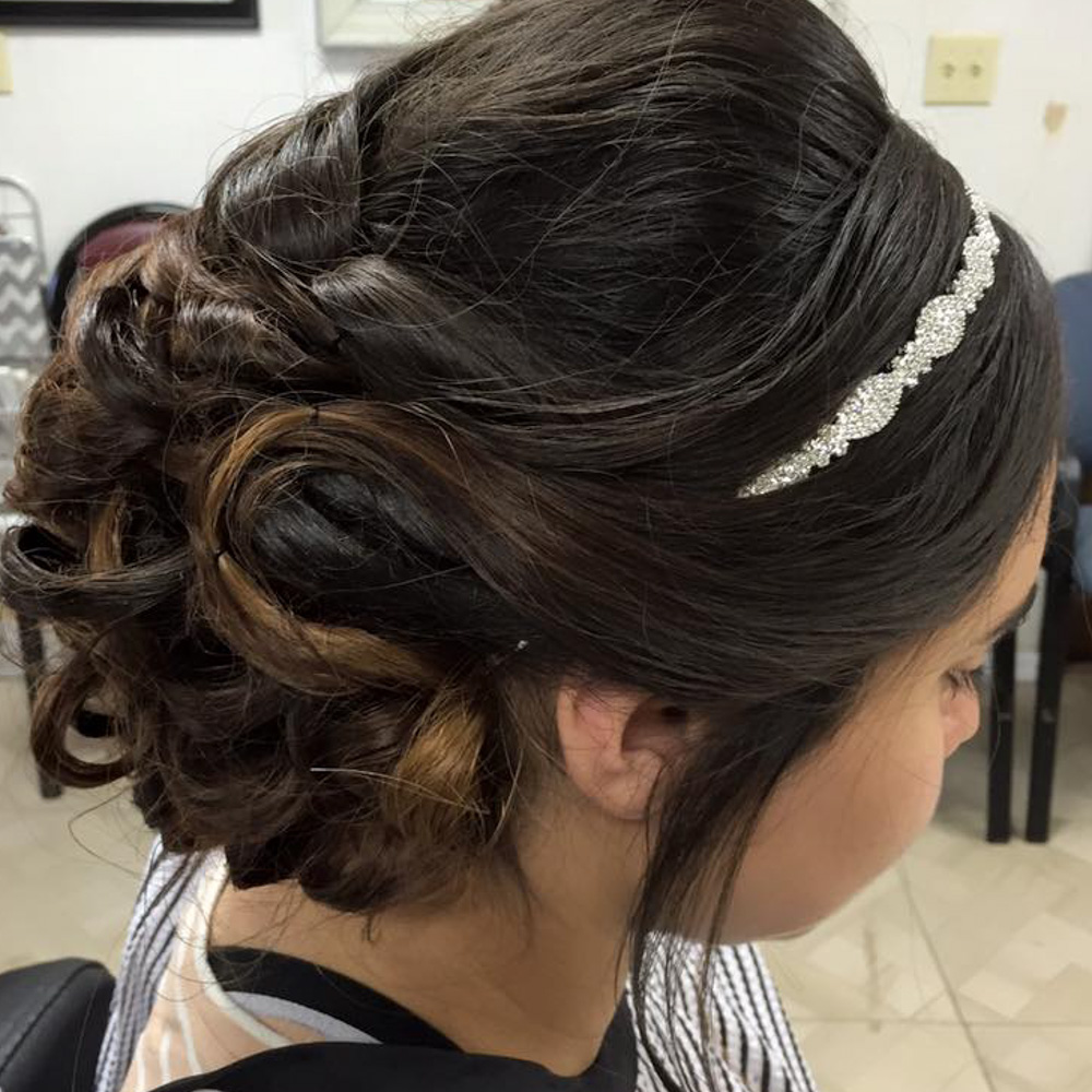 A side profile of a young woman with a voluminous, textured brunette updo consisting of soft curls and woven braids. The look is accessorized with a delicate, thin silver rhinestone headband and includes face-framing tendrils, perfect for a bridesmaid or formal gala.