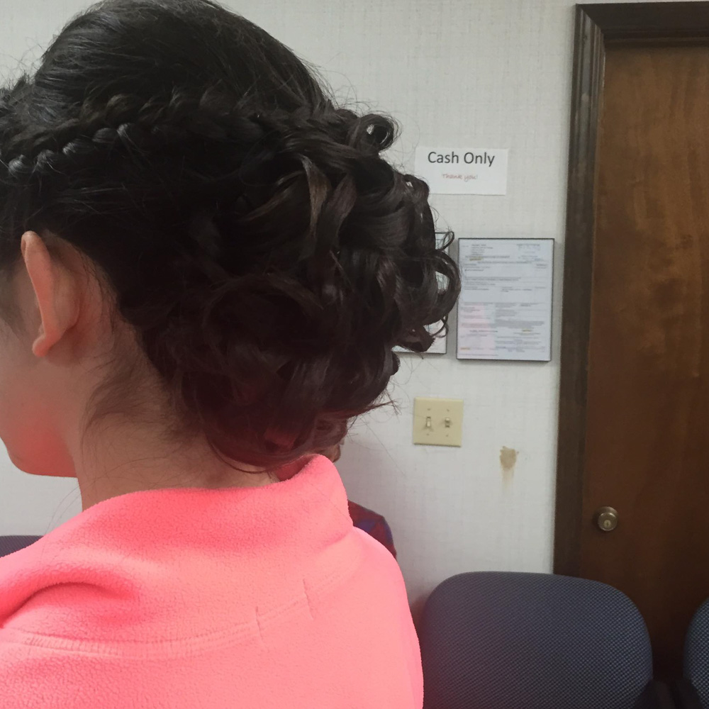 A back view of a dark-haired formal updo featuring a series of intricate, tight curls pinned into a voluminous low bun. A thin braid wraps around the side of the head to meet the curls, creating a romantic and secure hairstyle for a special event or dance.