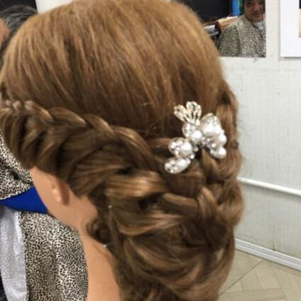 A rear view of a warm honey-blonde braided updo on a styling mannequin, featuring a thick crown braid that transitions into a low, textured bun. The hairstyle is accented with a decorative pearl and crystal floral hair clip, demonstrating formal hair-weaving techniques at Beauty Town Salons.